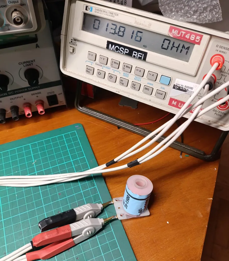 A Hewlett-Packard 3468A bench multimeter in 4-wire ohms mode reading 13.816 ohms, connected by four white leads and Kelvin clips to the lugs of a blue-labelled AE-26-1500 coil on a green cutting mat.