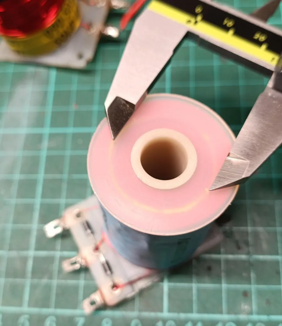 Top view of a dual-wound flipper coil with blue label and three lugs with diodes, with caliper jaws across the bobbin flange. A faint ring in the translucent pink flange shows where the inner power winding ends and the outer hold winding begins.
