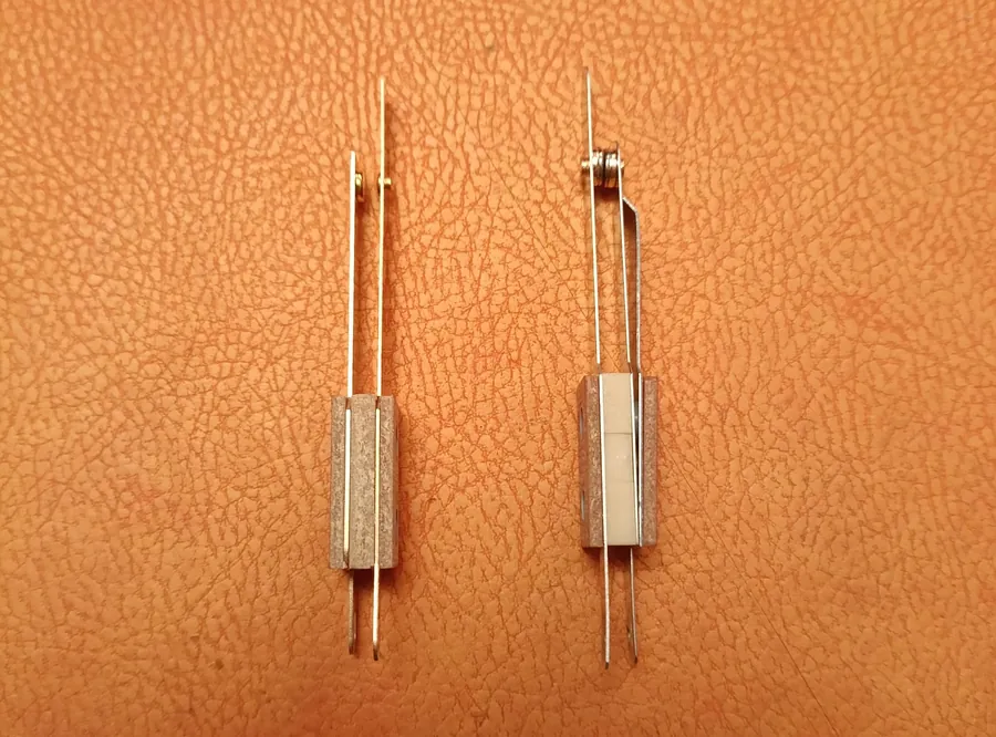 Two pinball leaf switches side by side against an orange background. Left: a normally-open switch with small gold-plated contact tips. Right: a normally-closed EOS-style switch with visibly larger bimetallic contact pads, the tungsten insert appearing as a distinct lighter-coloured rectangle against the steel blade.