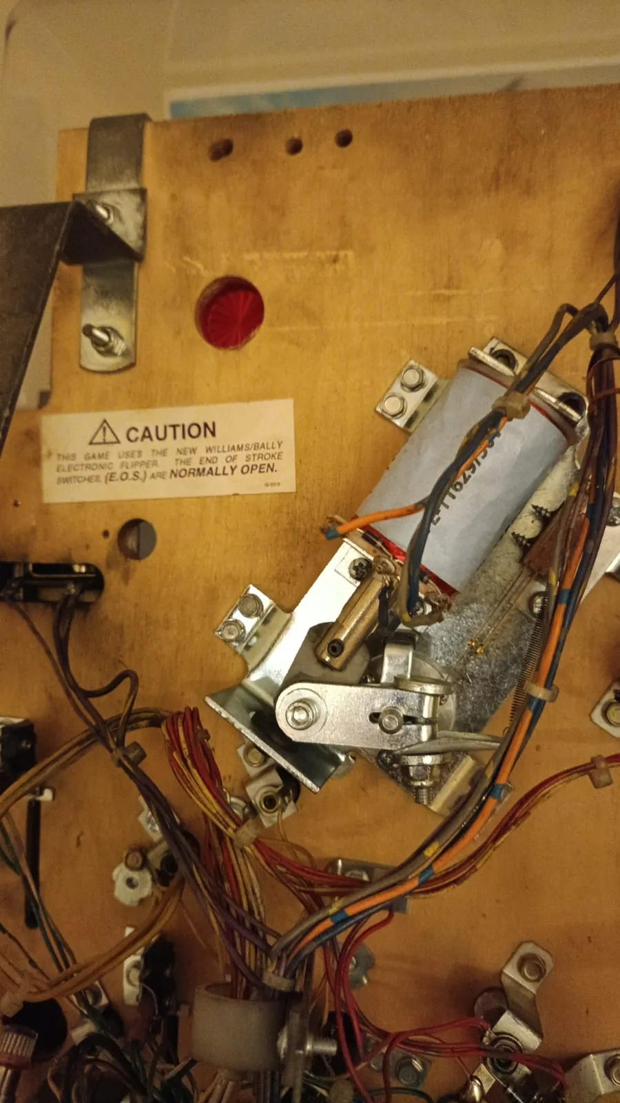 Underside of a Williams White Water playfield showing a Fliptronics II flipper mechanism. A printed caution label reads: "THIS GAME USES THE NEW WILLIAMS/BALLY ELECTRONIC FLIPPER. THE END OF STROKE SWITCHES (E.O.S.) ARE NORMALLY OPEN." The normally-open EOS switch blade and the dual-wound flipper coil are visible to the right of the label.