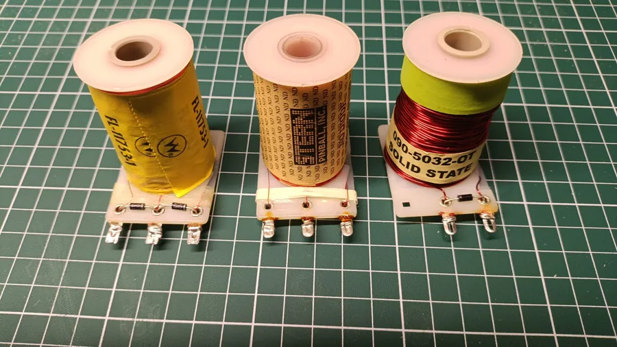 Three pinball flipper coils on a green cutting mat. Left: a Williams dual-wound coil (yellow label, FL-11753-1) with a flyback diode and three solder lugs. Centre: a Stern dual-wound coil (Stern Pinball Inc. label) with three lugs and no flyback diode. Right: a Data East single-wound coil (090-5032-OT Solid State label) with a flyback diode and two lugs.