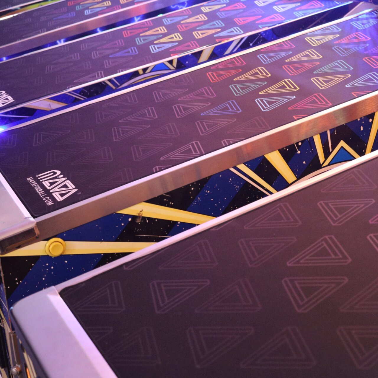 MAYA Glass Defender on a row of lit pinball machines, shot from above, printed pattern across the full width of the glass