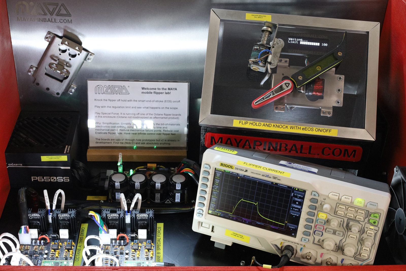 The full contents of the MAYA mobile flipper lab display cabinet with the glass removed, showing a PC power supply, MAYA power-conditioning capacitors, two Octane flipper driver boards, a Rigol oscilloscope displaying a flat-topped flipper current trace, and the flipper and knocker demo rig behind its own glass panel