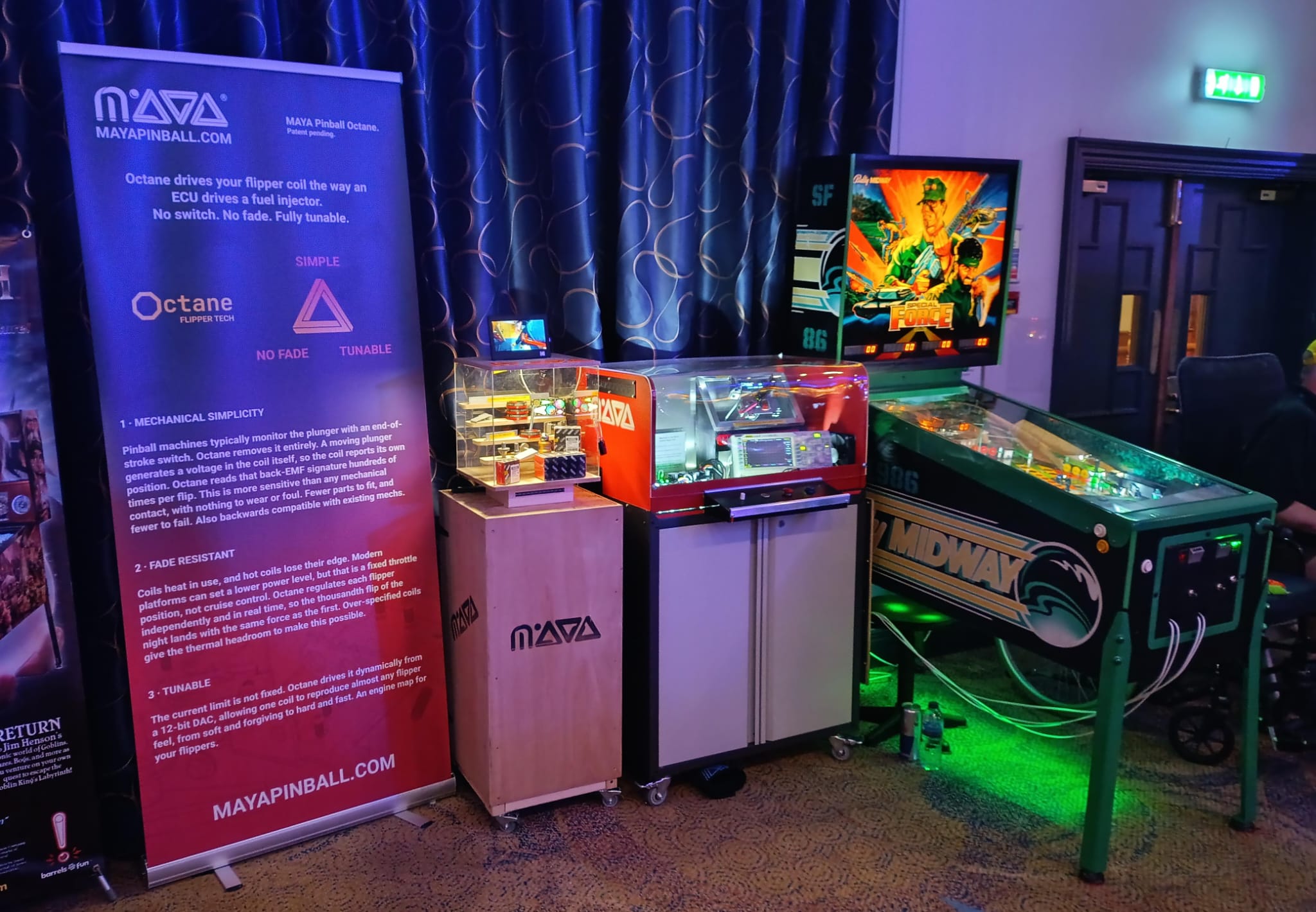 The MAYA Octane exhibit on the main hall floor at UK Pinfest 2026: a banner explaining the platform, a small display box of MAYA Pinball Amazon store items, an interactive demo enclosure behind clear acrylic, and an 80s Bally machine on the right with its original flippers replaced by the Octane system